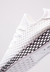 Vit sportskor med ett strukturerat nätöverdrag och svarta gallerdekoreringar på sulan, med vita snörningar och en rundad krage.