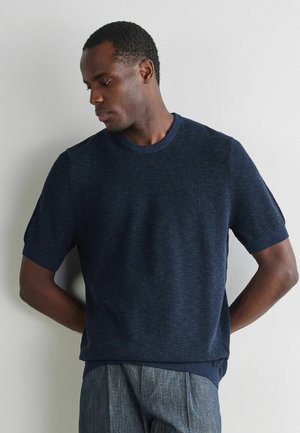 Mężczyzna w granatowej, krótkom rękawowej, fakturowanej koszulce z dzianiny oraz szarych spodniach stoi przy białej ścianie, patrząc w dół z rękami za plecami.