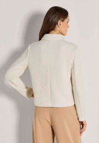 Frau mit braunen Haaren, die eine strukturierte weiße Langarmjacke und beige Hosen trägt, von hinten vor einem schlichten Hintergrund gezeigt.