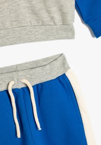 Grå kort sweatshirts med ribbad nederkant och blå ärmar, parat med blå joggingbyxor med grå midjeband och krämfärgad dragsko.