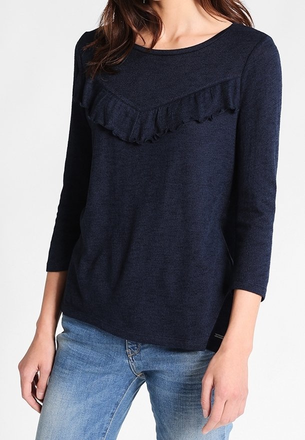 Femme portant un haut bleu marine à manches longues avec un détail de volants sur la poitrine, associée à un jean en denim bleu clair, debout devant un fond blanc.