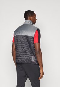 Muškarac nosi prsluk bez rukava u crno-sivoj kockastoj izvedbi preko crveno-crne majice kratkih rukava, stoji leđima okrenut prema gledištu na jednostavnoj pozadini.