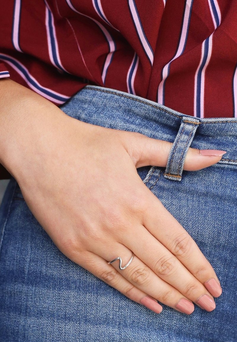 Zilveren golfvormige ring gedragen op een hand die rust op een blauwe denim tailleband, in contrast met een diep rode en gestreepte stoffen shirt.