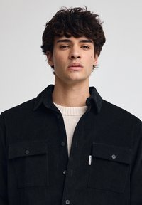 Czarna koszula z korduroy z zapięciem na guziki, z dwiema kieszeniami z przodu i wyprofilowanym kołnierzykiem. Pod spodem znajduje się kremowy sweter.