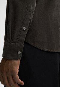 Camicia in corduroy scuro con tessuto testurizzato, caratterizzata da un polsino con bottoni e dettagli delle cuciture visibili, indossata sopra un indumento nero.