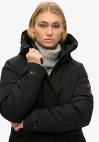 Zwarte pufferjack met een hoge kraag, ritssluiting en afneembare capuchon. Heeft een logopatch op de mouw en ribbelstructuur in de binnenkraag.