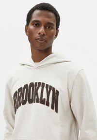 Sudadera beige clara con una textura suave, que presenta un texto bordado en marrón oscuro que dice "BROOKLYN" en el pecho y una capucha con cordones.
