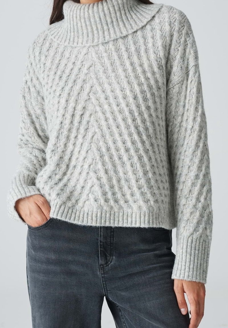 Femme portant un pull à col roulé en maille torsadée gris clair avec poignets côtelés et jean gris foncé, main dans la poche, sur un fond uni.