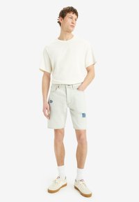 Pantalones cortos de mezclilla azul claro con detalles de patchwork, combinados con una camiseta blanca de manga corta y zapatillas crema sobre calcetines blancos.