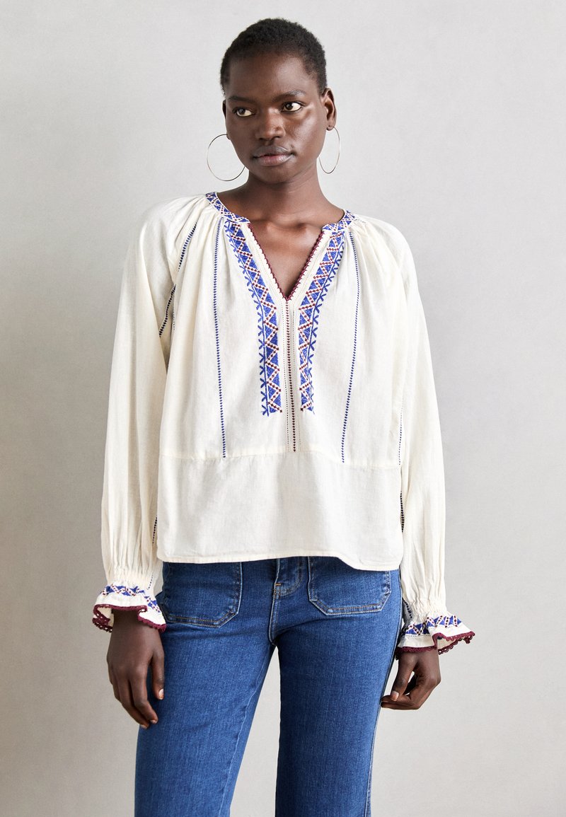 Blouse à manches longues de couleur crème, ornée de broderies bleues et rouges le long de l'encolure et des poignets ; coupe décontractée avec manches froncées.