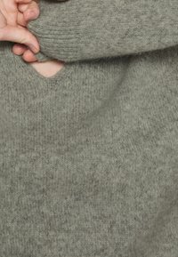 Main saisissant la manche d'un pull gris tricoté près d'un petit trou au niveau du poignet.