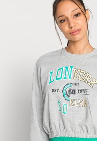 Sudadera gris de manga larga con estampado de texto en verde y beige. Tiene un cuello redondo y dobladillo elástico, hecha de un material suave.