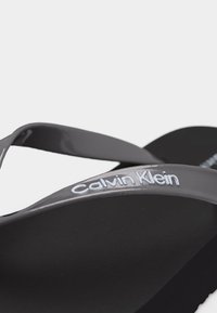 Μαύρο σανδάλι τύπου σαγιονάρα με σκούρο γκρι λουράκι που φέρει το λευκό λογότυπο «Calvin Klein» στο πλάι.