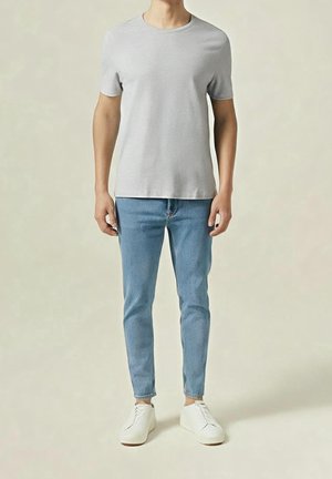 Person trägt ein hellgraues T-Shirt mit kurzen Ärmeln, hellblaue Slim-Fit-Jeans und weiße Turnschuhe vor einem einfarbigen Hintergrund.