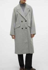 Cappotto grigio in misto lana con design doppiopetto, ampi rever, due tasche frontali e vestibilità rilassata. Indossato sopra un dolcevita.