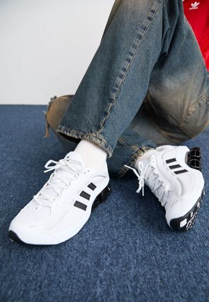 Personne portant des chaussures de sport blanches et noires, des chaussettes blanches et un jean bleu, assise sur un sol recouvert de moquette bleue.