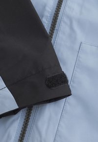 Mėlyna ir juoda striukė su juodu rankogaliu, kuriame yra Velcro uždarymas, ir kontrastinga juoda užtrauktuku priekyje.