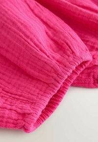 Tissu texturé rose vif avec des bords élastiques froncés, probablement une partie d'une manche ou d'une jambe de pantalon, présenté sur un fond clair.
