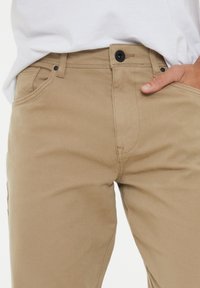 Beige Baumwollhosen mit glatter Textur, standardmäßiger Fünf-Taschen-Design, mit schwarzem Knopfverschluss und Gürtelschlaufen.