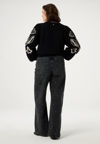 Zwarte gebreide trui met decoratieve borduursels op de opgezette mouwen, gecombineerd met donkere jeans met versieringen en een uitlopende snit.
