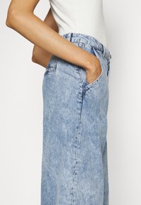 Personne portant un jean en denim bleu clair, les mains dans les poches, et un haut blanc sans manches sur un fond uni.