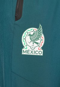 Türkiz sportzakó sima textúrával, amely egy kör alakú logót ábrázol. A logó szárnyas mintázatot és vastag betűkkel a "MÉXICO" szót mutatja.