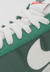 Green sneaker with white details, textured laces, and a white logo. Features a smooth fabric upper and rubber sole.