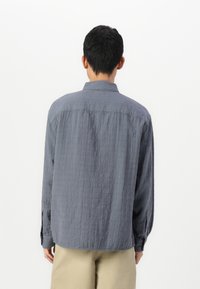Camisa gris de manga larga hecha de tela texturizada, con cuello y puños abotonados. La vista trasera muestra un corte relajado con un dobladillo alargado.