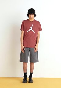 Rød bomulds T-shirt med et hvidt Jordan-logo, parret med grå shorts og sorte sko. Enkel design, lige snit, korte ærmer.