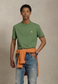 Mladenič stoji z eno roko v žepu, oblečen v zeleno majico, modre kavbojke, oranžno jopico, zavezano okoli pasu, in s uri na zapestju.