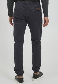 Navy jogger pants with elastic waistband and cuffs, featuring a smooth texture and two back pockets, accented with a small brown label.
