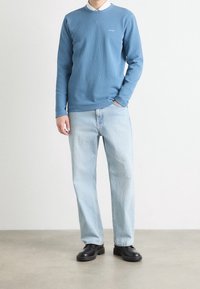 Helles blaues Langarmshirt mit Rundhalsausschnitt und dezentem Logo, kombiniert mit hellblauen Jeans und schwarzen Schuhen. Glatter Stoff und lässige Passform.