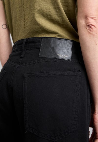 Black denim jeans featuring a textured leather patch with embossed logo, side pockets, and a smooth finish. Olive green shirt visible above.