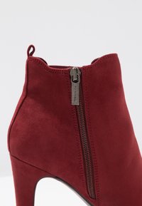 Red suede ankle boot with a high heel, featuring a side zipper and smooth texture. The design includes stitched details along the seams.