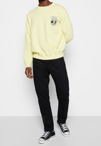 Gelbes Sweatshirt mit rundem Ausschnitt und elastischen Bündchen, mit einem grafischen Aufnäher auf der Brust. Kombiniert mit schwarzen Hosen und schwarzen Turnschuhen.