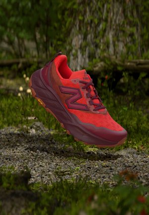 Czerwono-bordowy but sportowy unoszący się nad żwirową ścieżką na leśnej polanie, z grubą podeszwą z logo Vibram i siateczkową cholewką.