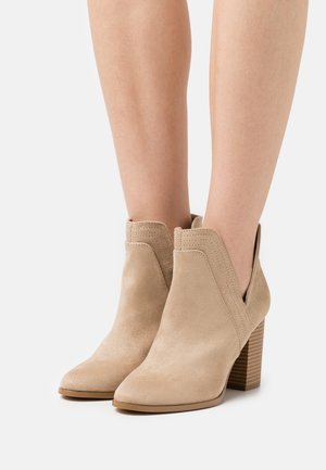 Beige Wildleder-Ankle Boots mit spitzem Zehenbereich, eingeschnittenen Seitenöffnungen und einem gestapelten Absatz mit horizontalen Rillen.