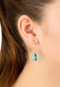 Los pendientes colgantes en tono dorado presentan una piedra preciosa azul en forma de lágrima con una superficie facetada, colgando de un delgado gancho de alambre.