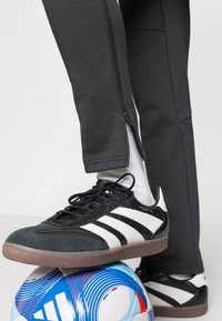 Αθλητικά παπούτσια ασπρόμαυρα με γκρι σουέντ λεπτομέρειες πάνω σε μια ποδοσφαιρική μπάλα με μπλε και λευκό σχέδιο. Γκρι παντελόνι με ορατά φερμουάρ στα πλάγια.