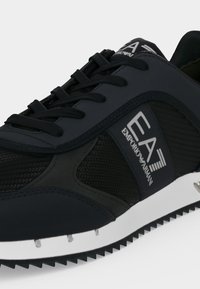 Μαύρο και σκούρο γκρι αθλητικό παπούτσι EA7 Emporio Armani με μαύρα κορδόνια, υφασμάτινες πάνελ με υφή και λευκή σόλα με αυλακώσεις.