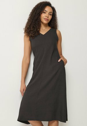 Frau mit lockigem Haar, die ein ärmelloses schwarzes Midikleid mit V-Ausschnitt trägt, vor einem einfarbigen hellen Hintergrund stehend, Hand in der Tasche.