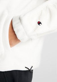 Ręka w kieszeni białej polarowej kurtki z czarnym regulowanym sznurkiem i małym czarno-czerwonym logo na rękawie.