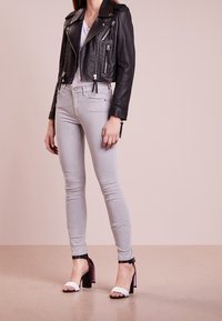 Chaqueta de moto de cuero negro con cremalleras plateadas, combinada con jeans ajustados gris claro y sandalias de tacón blanco con detalles en negro.