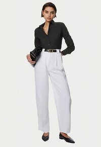 Black button-up shirt, high-waisted white pleated pants, black belt, and pointed black shoes. Model holding a black handbag.