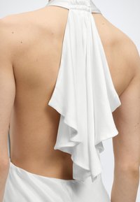 Πίσω όψη ενός ατόμου που φοράει λευκό φόρεμα με λουράκι στο λαιμό και βολάν στο κέντρο της ανοιχτής πλάτης.