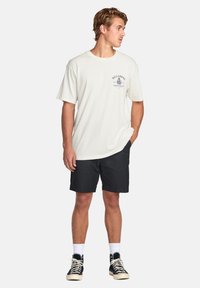 Billabong grafisk T-skjorte i off-white bomull, med en blå logo og design på brystet, kombinert med svarte shorts og høyhælte joggesko.