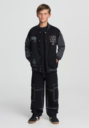 Giacca varsity nera con maniche grigie, con testo ricamato e una grafica. Abbinata a pantaloni cargo neri e scarpe nere con dettagli bianchi.