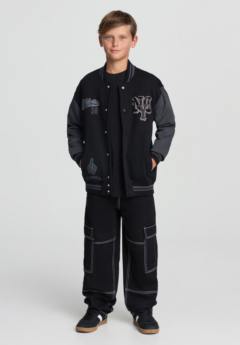 Czarna kurtka varsity z szarymi rękawami, z haftowanym tekstem i grafiką. Połączona z czarnymi spodniami cargo i czarnymi butami z białymi akcentami.