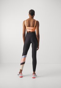 Oranssi urheil bra, jossa on säädettävät hihnat ja logodetail, yhdistettynä mustiin legginsseihin, joissa on värikäs geometrinen kuvio yhdellä jalalla.