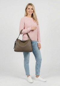 Sieviete rozā džemperī, gaiši zilās džinsās un baltās sporta kurpēs, kas tur lielu brūnu ādas somu uz kreisās rokas un smaida.